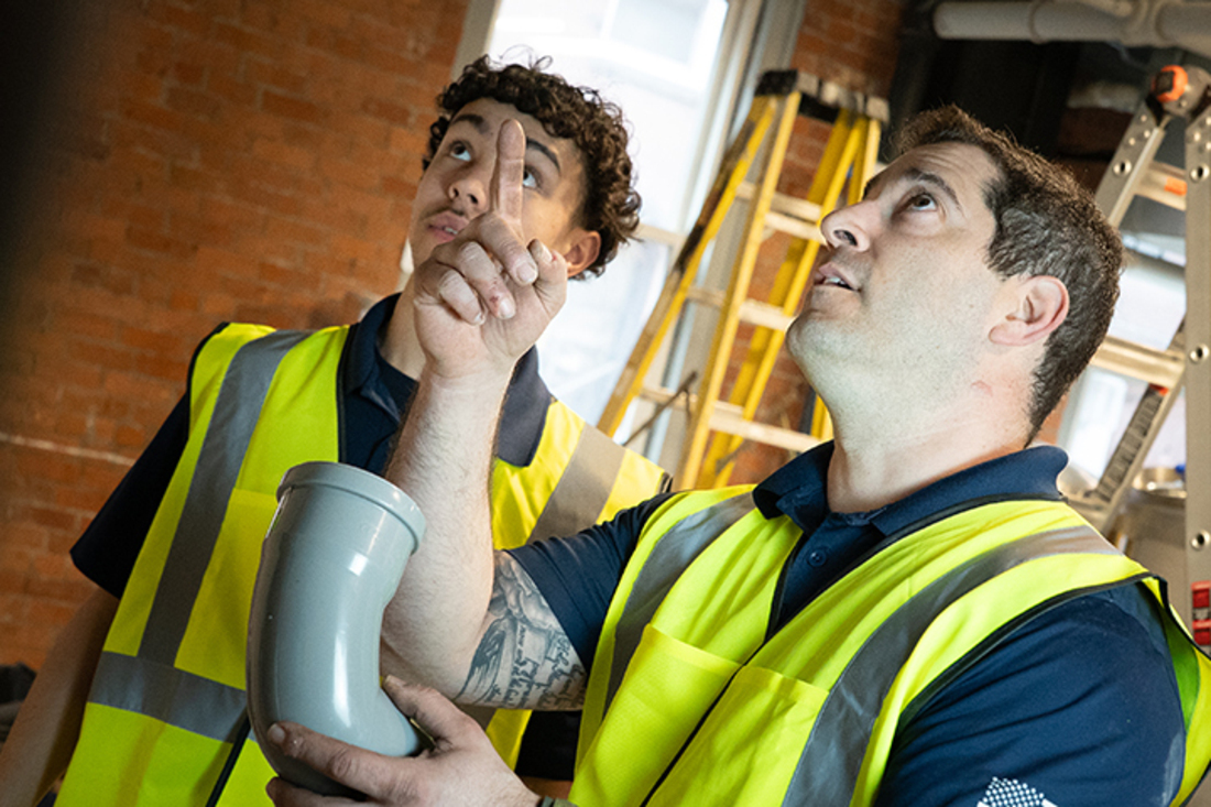 Two workers in high-visibility vests examining a gray innoflue elbow fitting inside a construction or mechanical room.