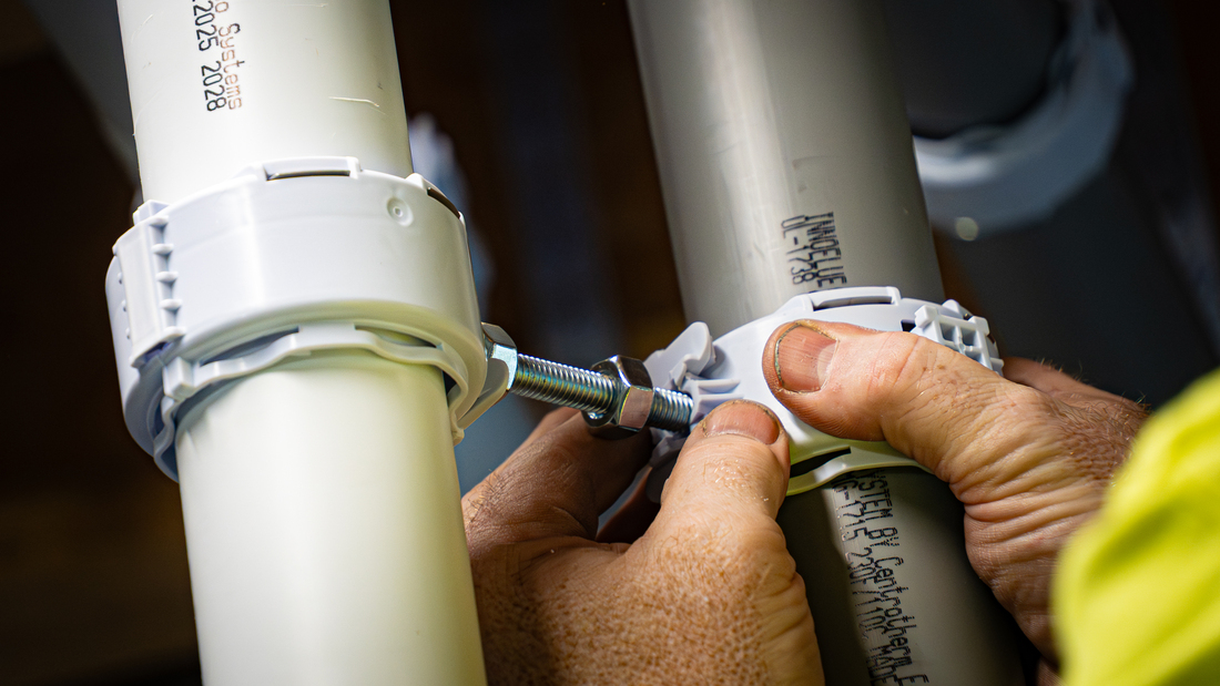 Close-up of hands fastening an octoclamp between white and gray pipes using a threaded bolt and nuts.