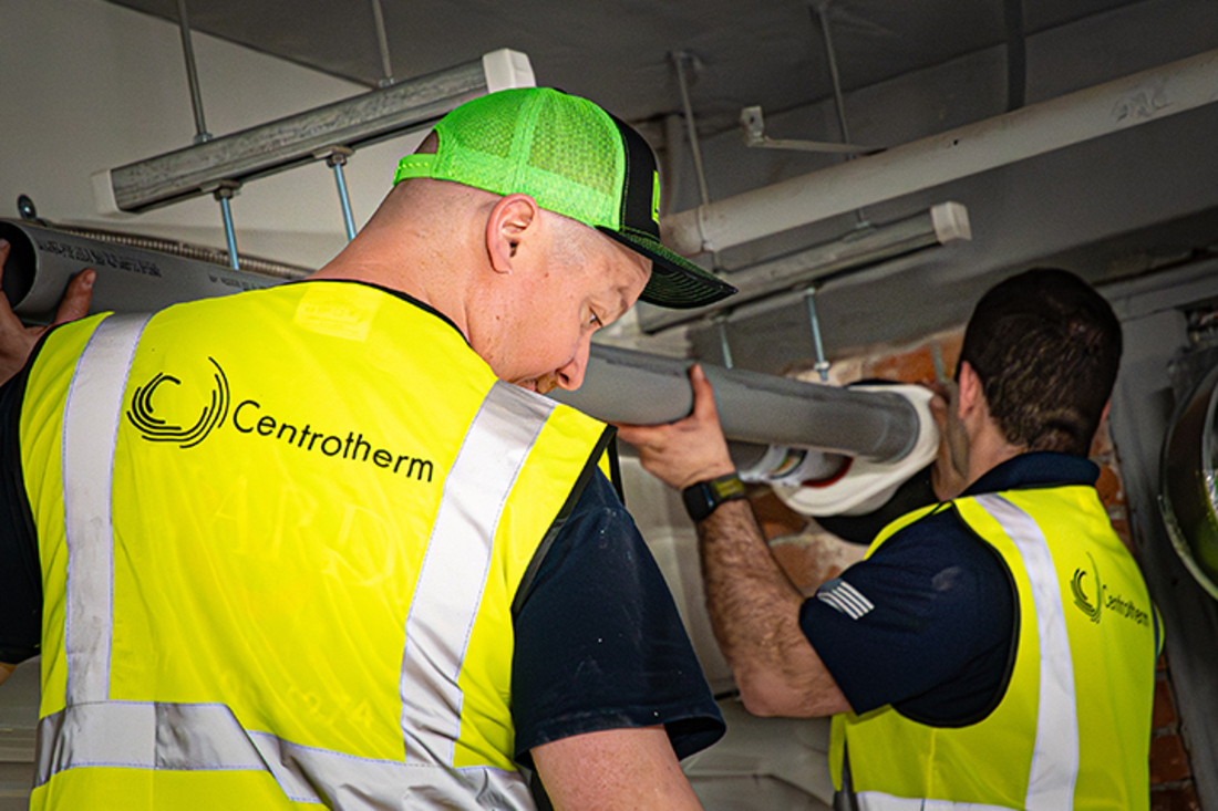 Two installers fitting gray innoflue pipe into a suspended system inside a commercial or industrial space.